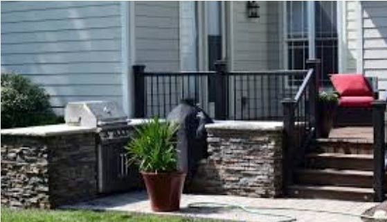 Front porch with stone posts, black railing, potted plant, grill, and red chair beside house steps
