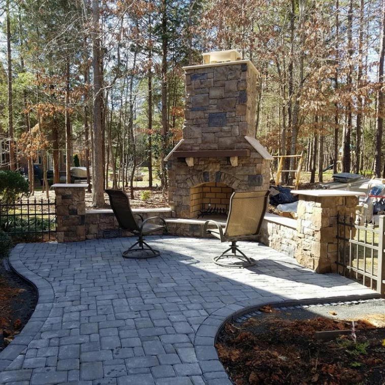Stone patio with outdoor fireplace and two chairs in a wooded backyard