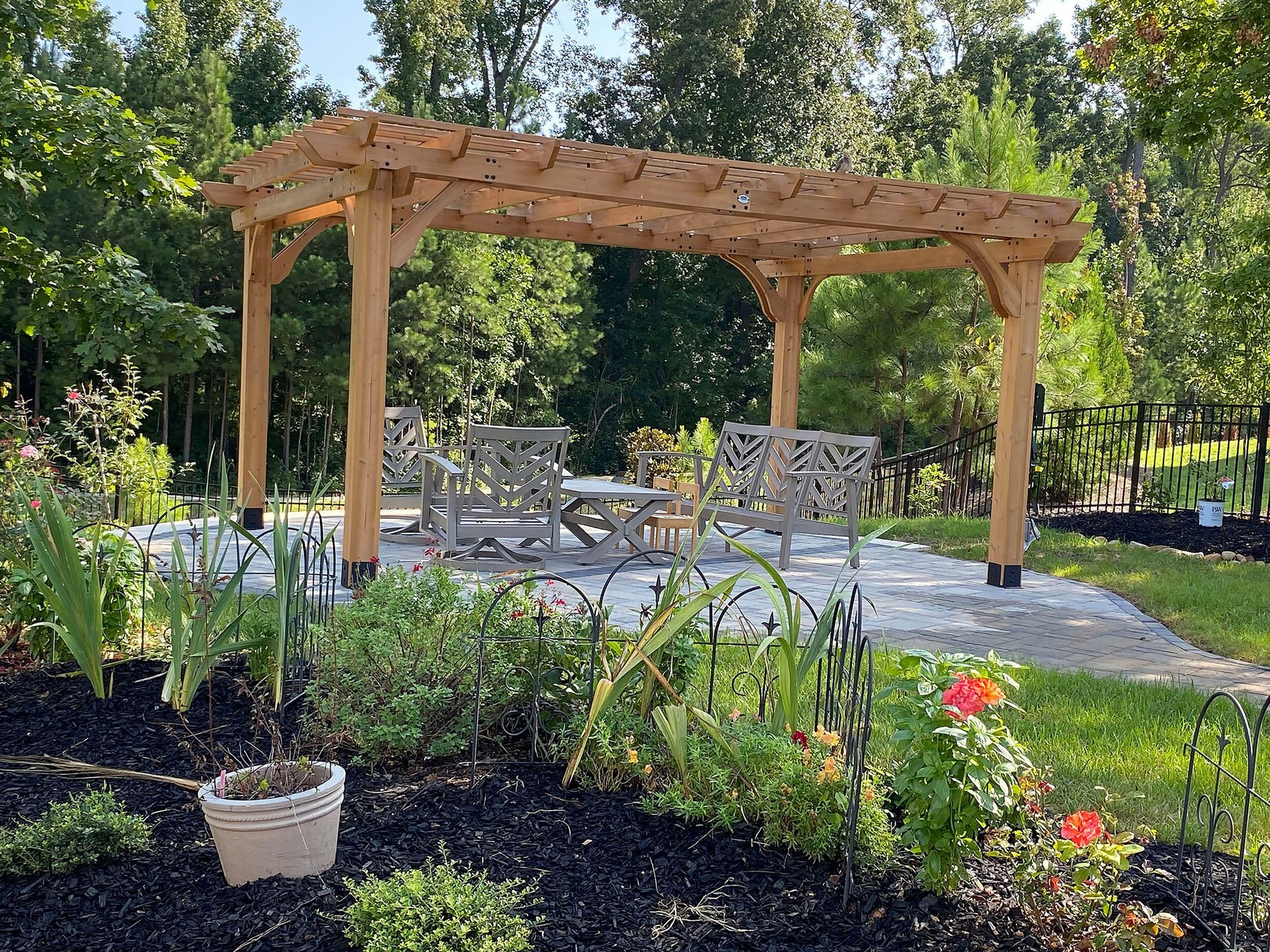 Wooden pergola over patio seating in a garden, with flowers and trees around it.