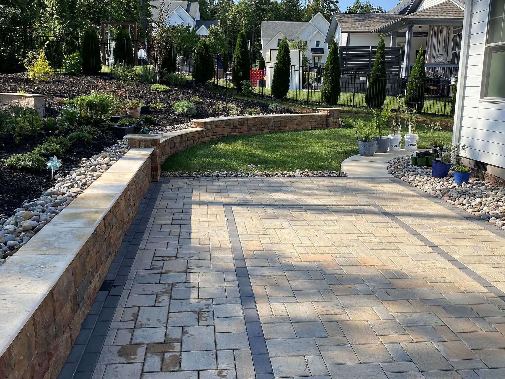 Backyard patio with stone pavers, curved retaining wall, garden beds, and lawn in sunlight