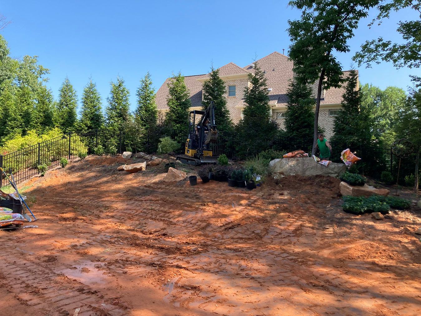 Bare dirt yard with a small stone retaining wall, trees, and a house in the background under a clear blue sky