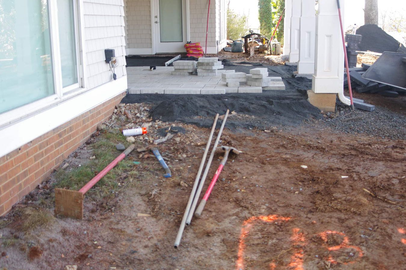 Front porch construction site with dirt, tools, and paving materials beside a house