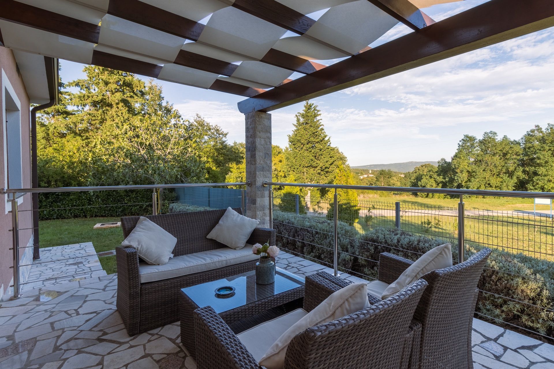 Covered patio with wicker seating, glass table, and a view of green fields and trees