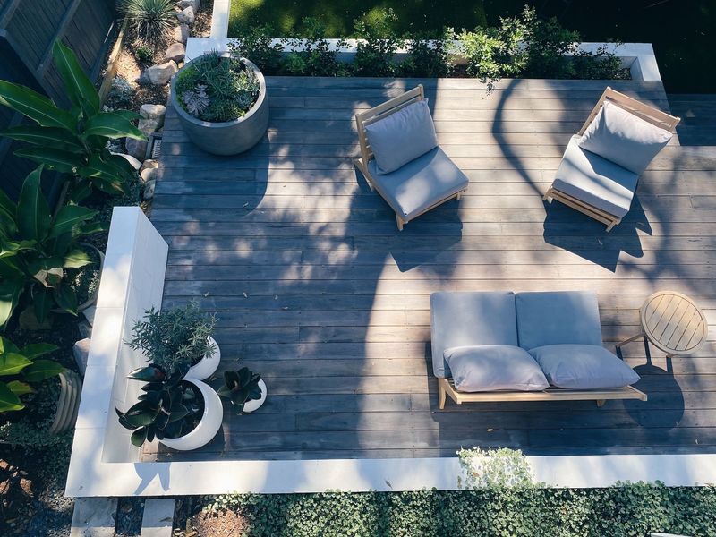 Patio with wicker chairs and cushioned bench on a sunlit wooden deck, surrounded by potted plants and greenery