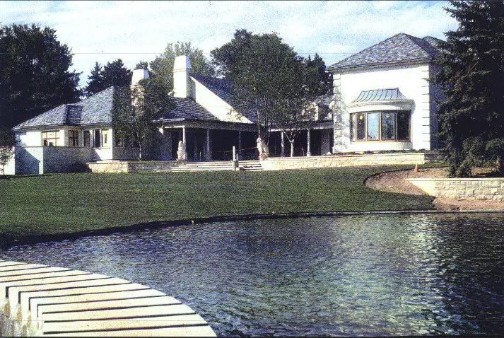 Large house beside a pond with a grassy lawn and trees, viewed from a wooden dock