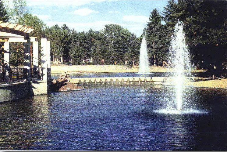 Park pond with fountains spraying water beside a shaded walkway and trees