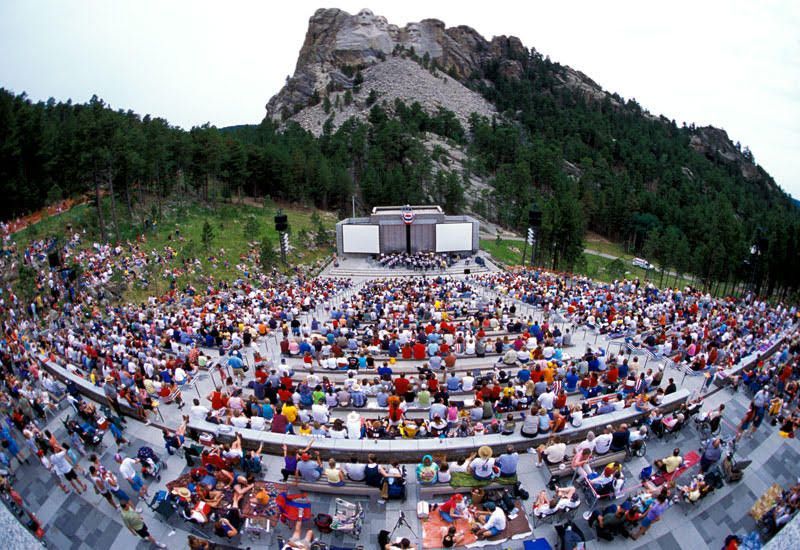 Outdoor concert crowd seated in a mountain amphitheater facing a stage, surrounded by trees and rocky cliffs