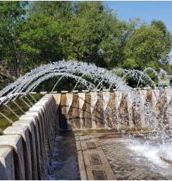 Water fountain arches spraying over a stone walkway beside trees and blue sky