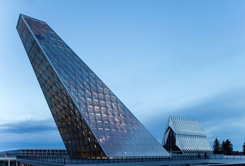 Modern glass pyramid building at dusk with a reflective facade and a small angular structure nearby