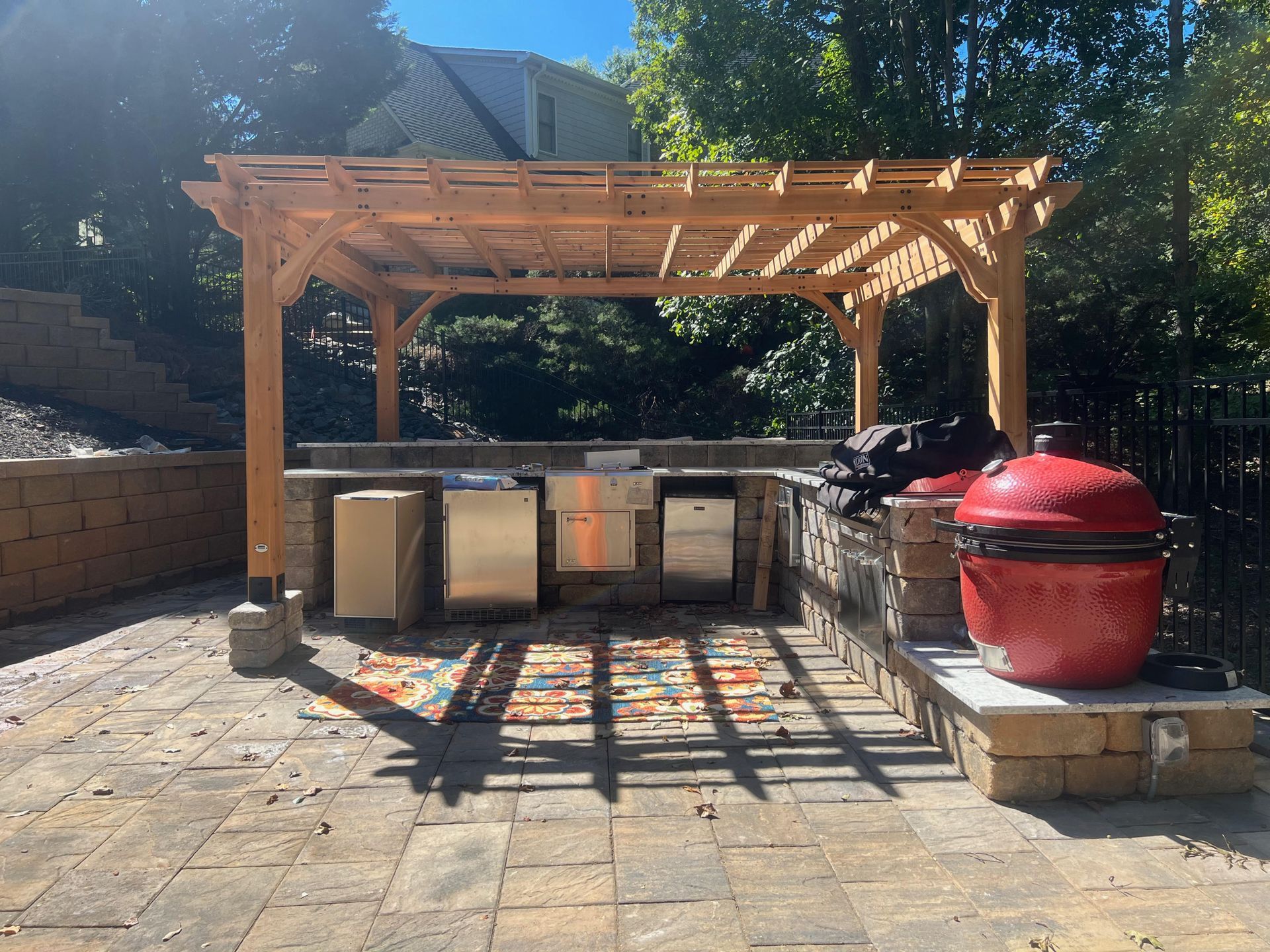 Backyard patio with a wooden pergola, grill, red smoker, and storage cabinets on stone paving