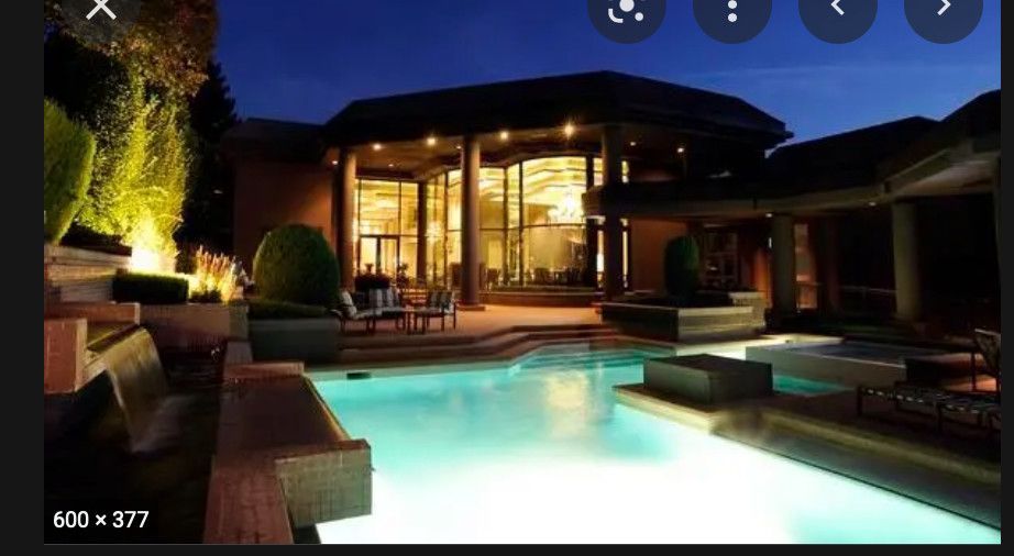 Luxury backyard pool at night with glowing water, patio seating, and a modern house in the background
