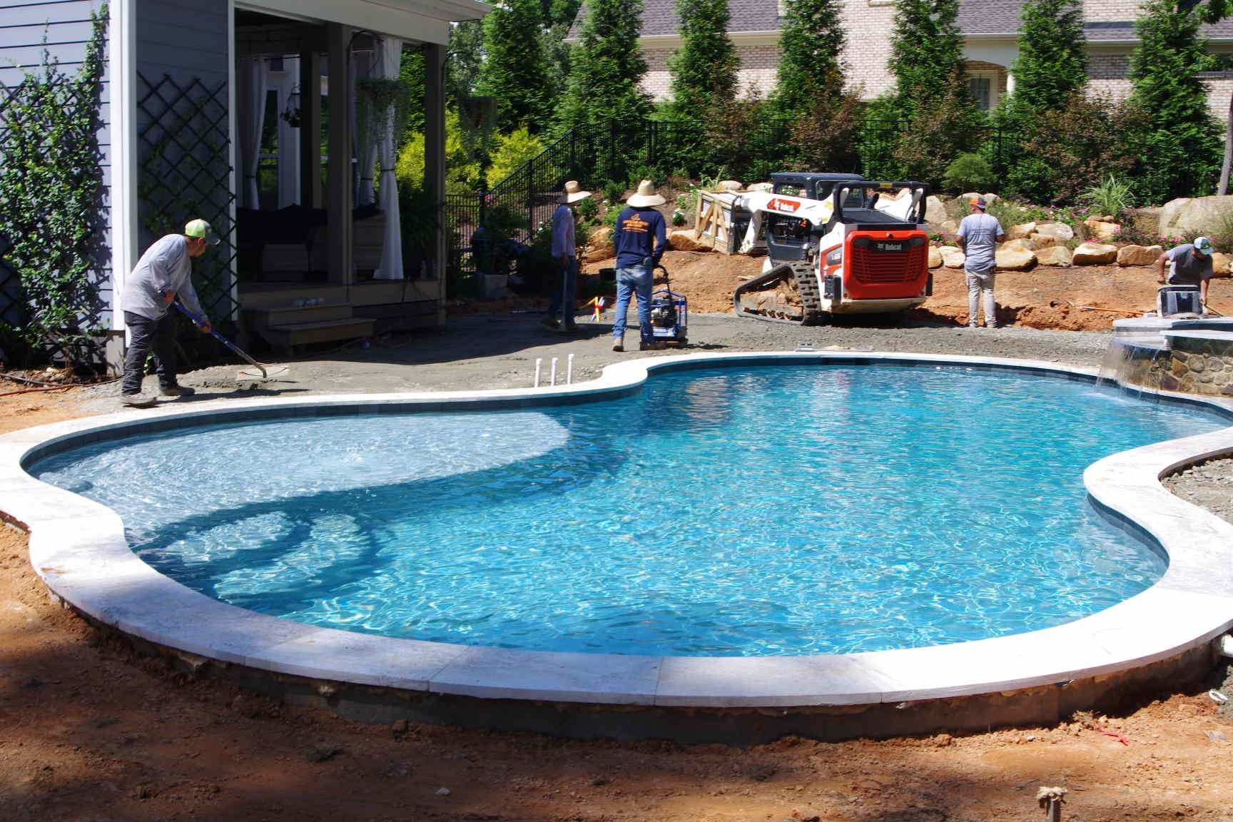 Backyard swimming pool under construction with workers, tools, and landscaping around it