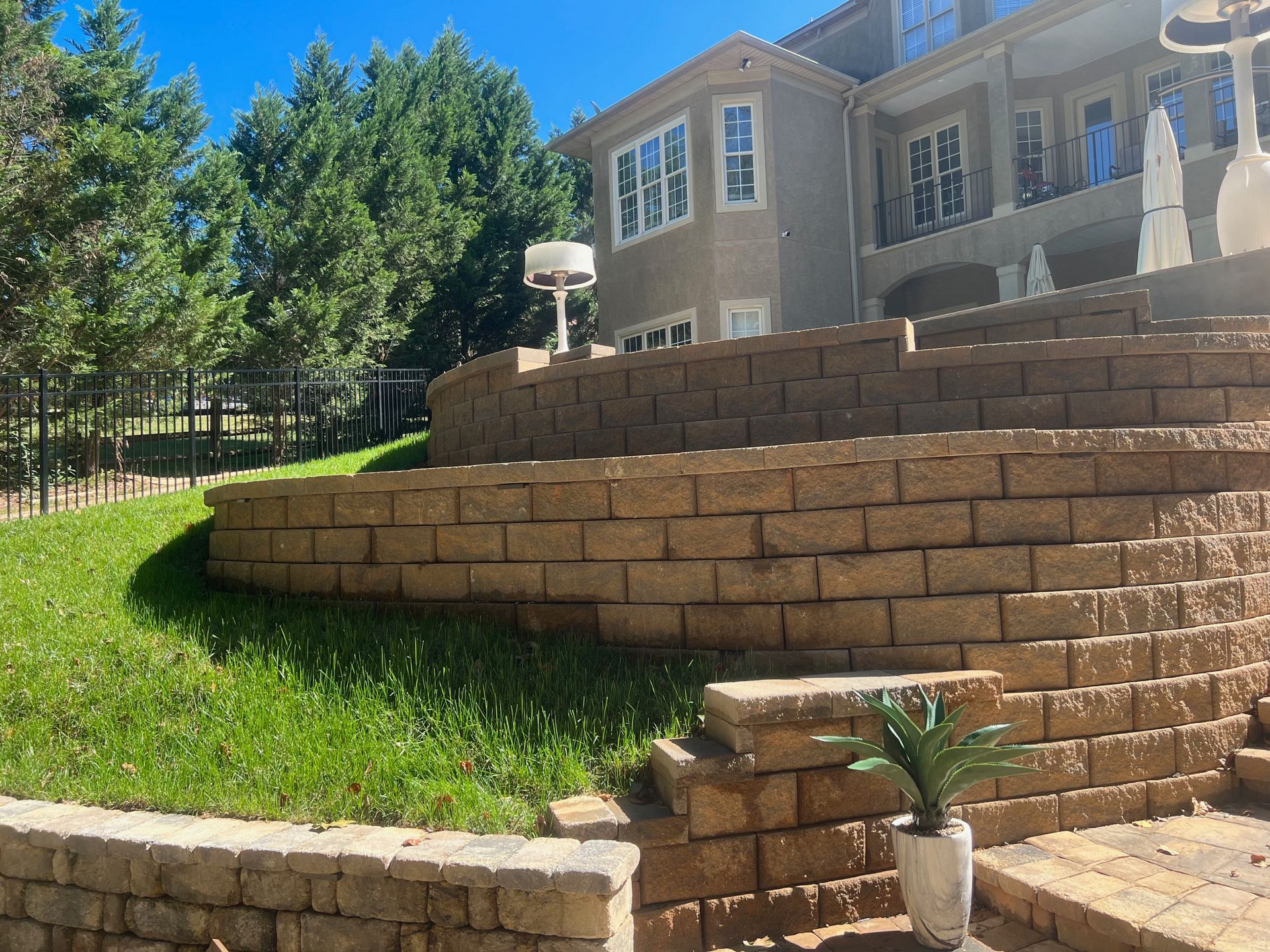 Stone terraced garden with brick retaining walls, green grass, potted plant, and a house with windows in the background