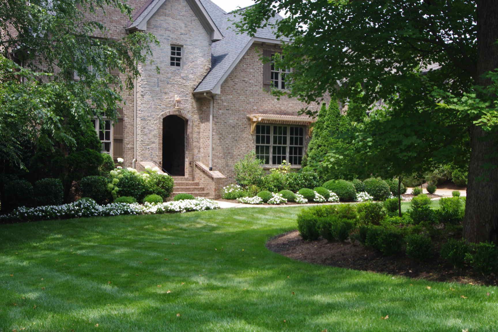 Stone house with arched doorway, lush green lawn, shrubs, and a shaded garden path