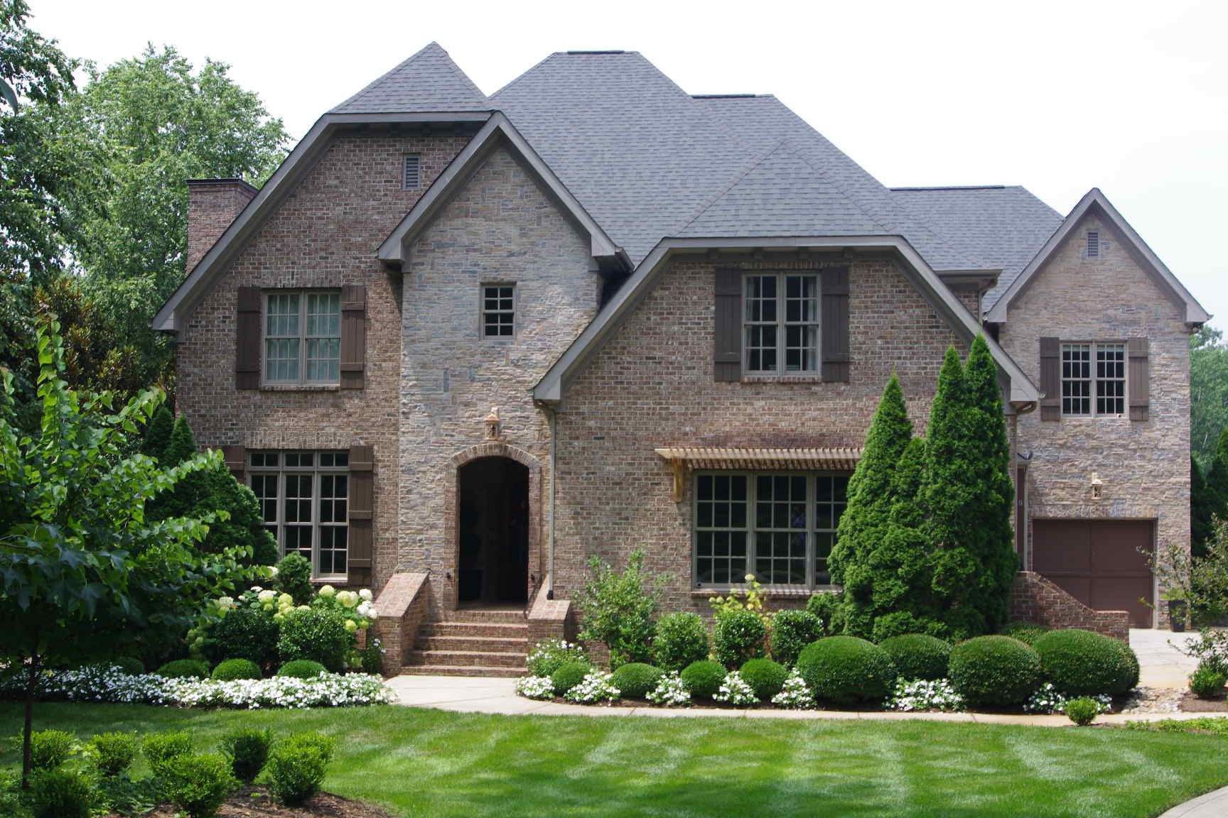 Large brick suburban house with gray roof, front steps, manicured lawn, and trees.