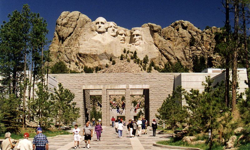 Tourists walking toward Mount Rushmore's entrance with the carved presidents visible above the trees