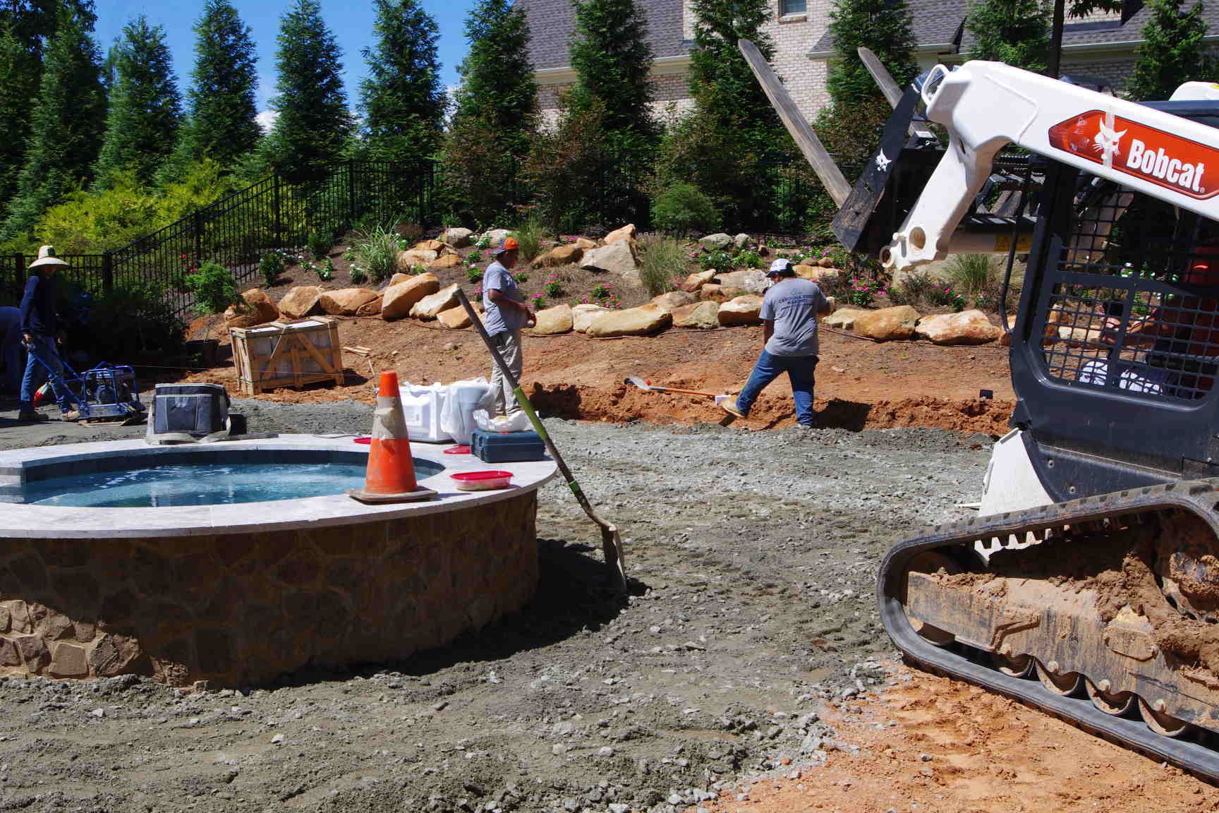 Construction crew grading gravel around a round pool, with an excavator and trees in the background
