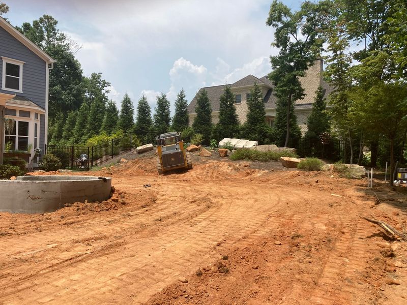 Backyard under construction with dirt grading, retaining walls, and trees beside a house