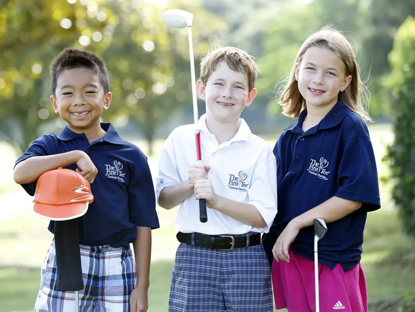 Three children in school uniforms holding golf clubs on a sunny outdoor course.
