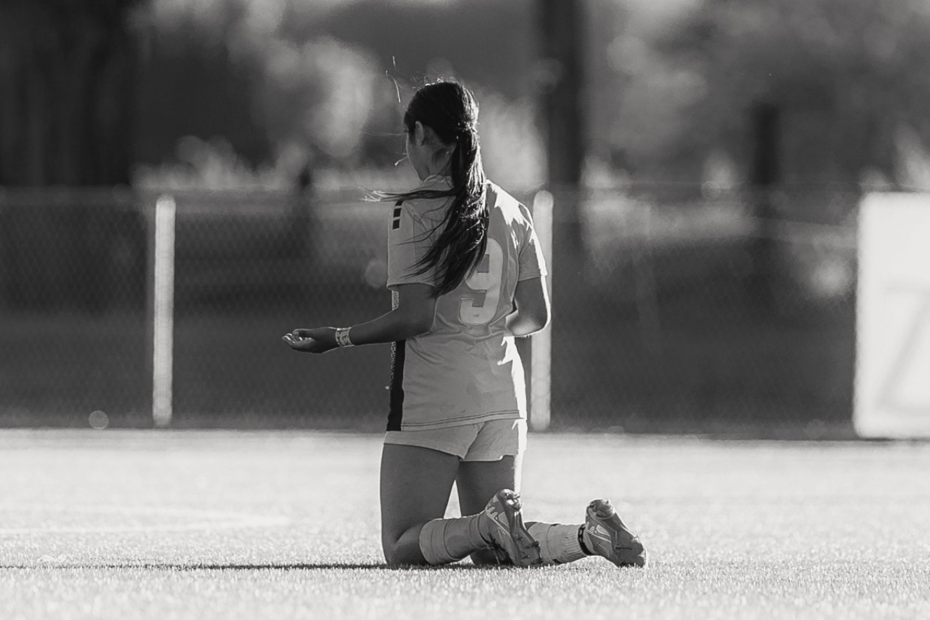 Kneeling soccer player on a field, holding a ball near the goal area.