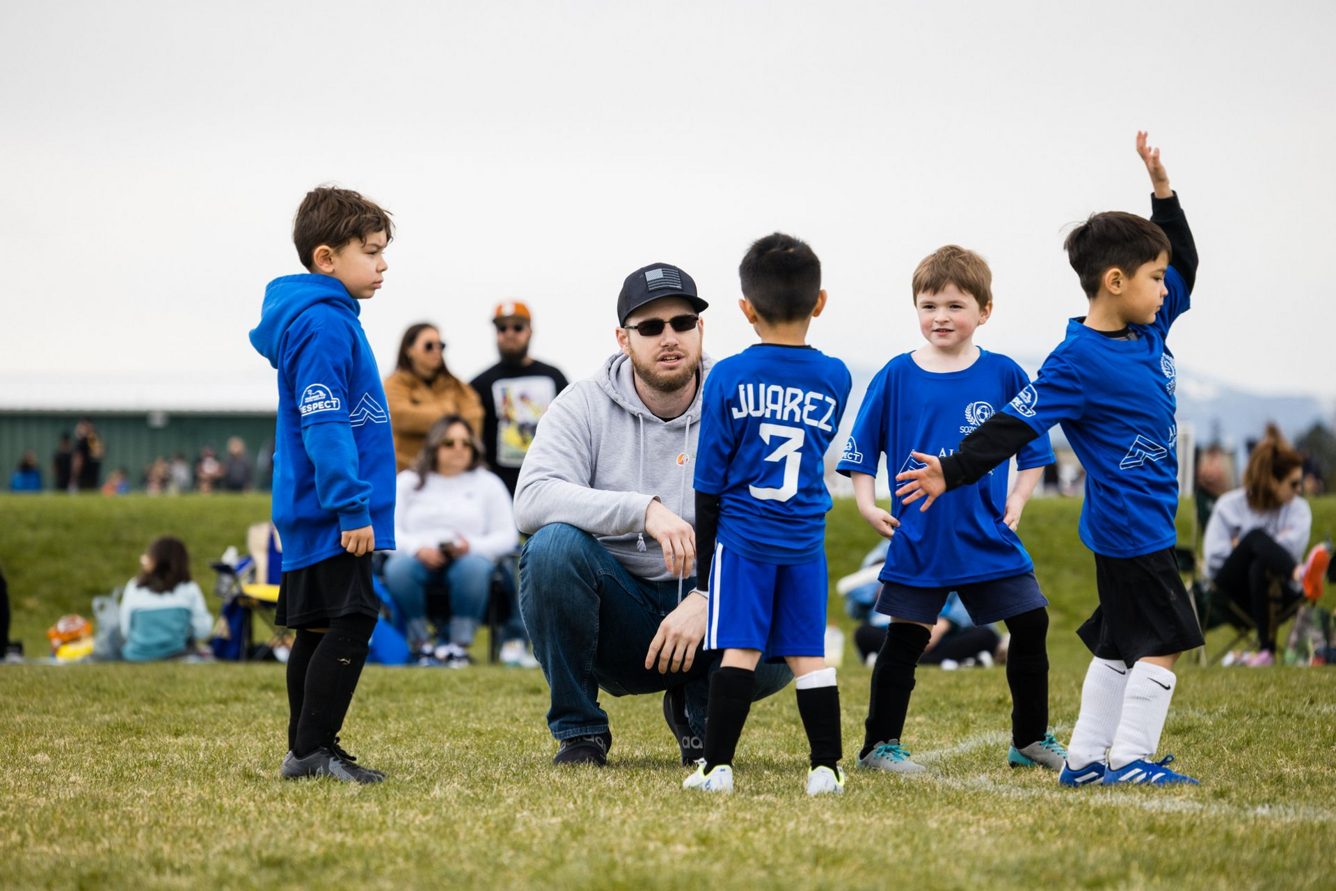 Children in blue soccer uniforms huddle on a grassy field during a game, with spectators in the background