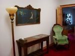 A floor lamp stands next to a wooden table and a green upholstered chair in a room with a dark painting on the wall.