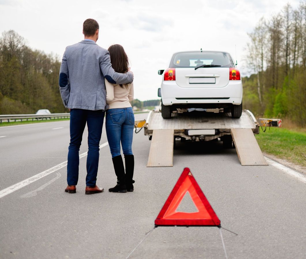 Una coppia si trova accanto a un'auto bianca in panne su una rampa di traino, con un triangolo rosso di segnalazione sul ciglio della strada.