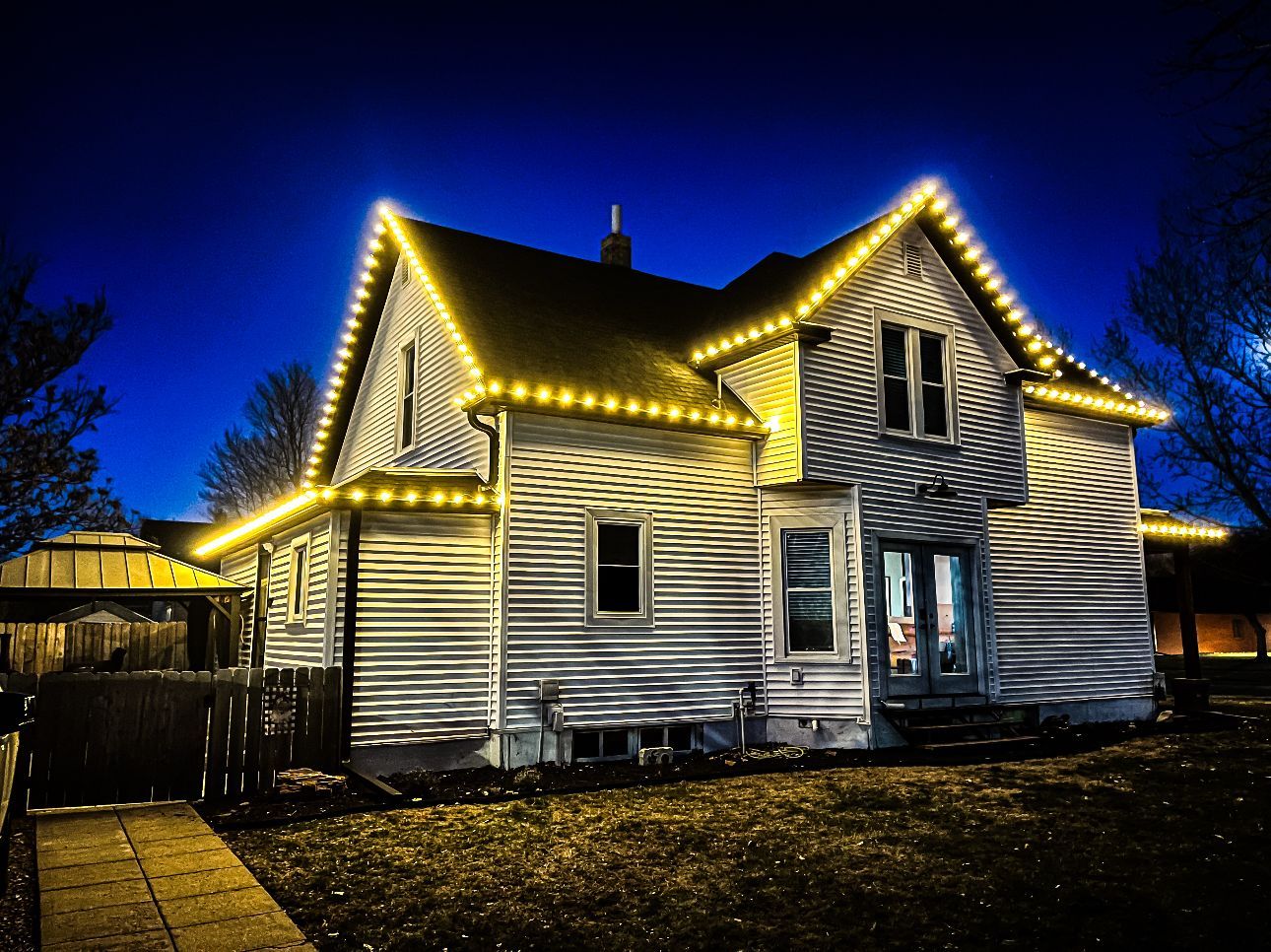 House at night, decorated with yellow Christmas lights along the roofline. Dark blue sky.
