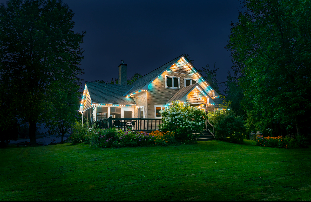 A cozy house at night, lit with colorful lights. Lush green grass and trees surround it.