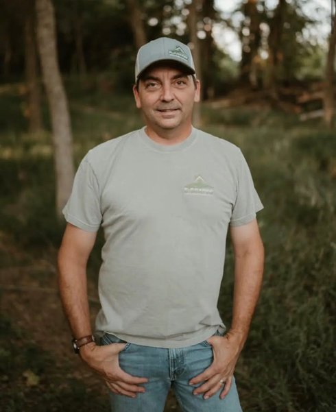 Man in gray shirt and cap smiles in front of red and yellow plants.