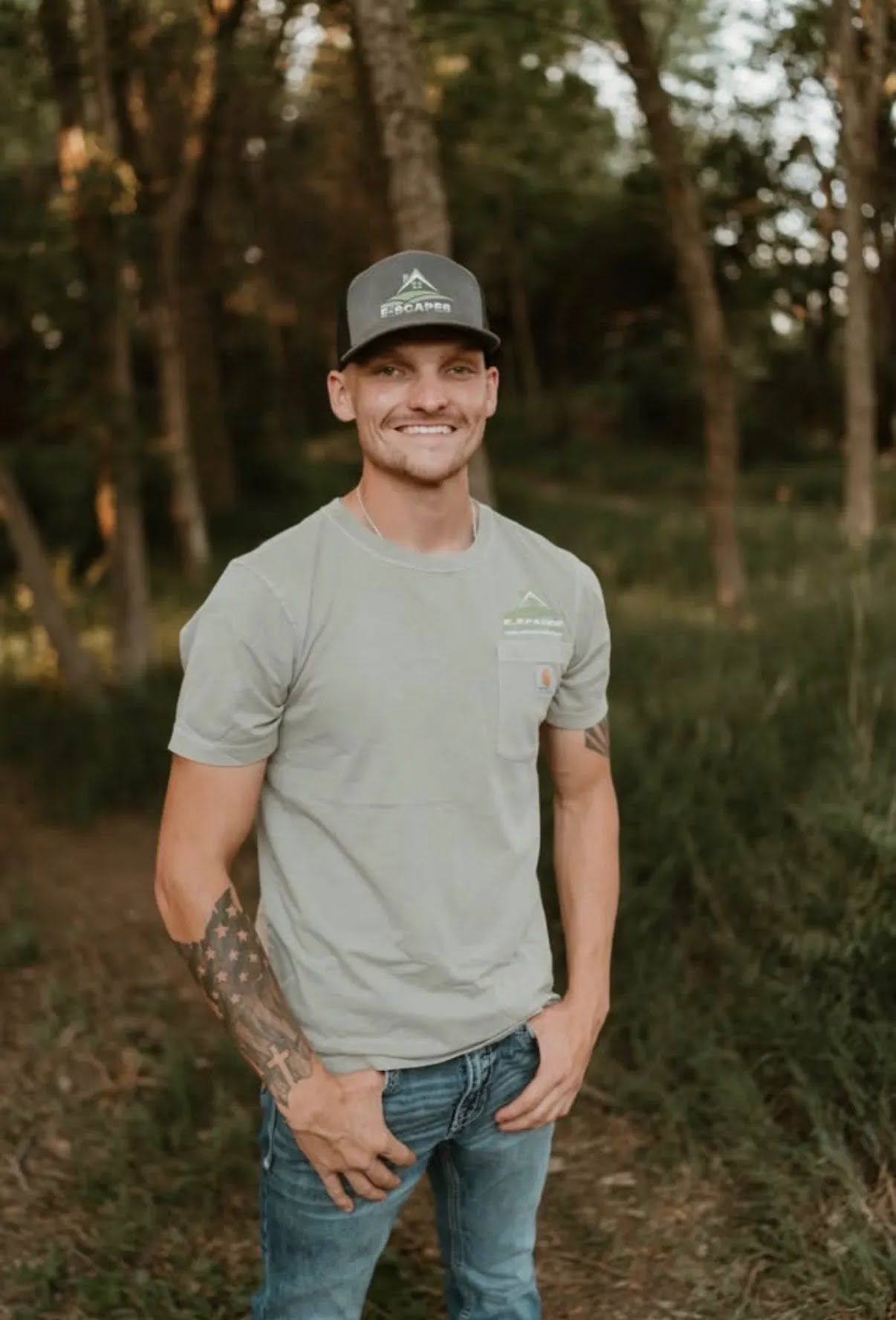 Man in white shirt smiles, hands in pockets, in front of a brick wall. He has tattoos on his arm.
