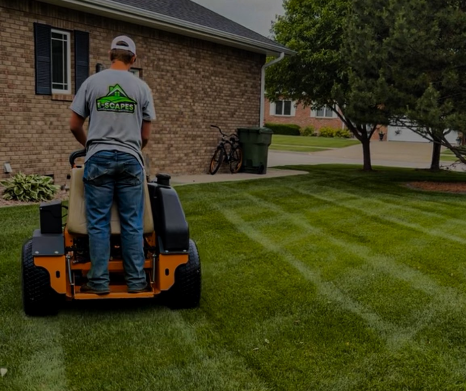 A man mowing a lawn with a riding mower, creating stripes. Brick house in the background.