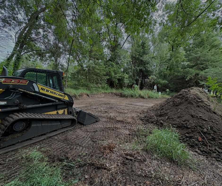 A New Holland skid steer at a construction site. The machine sits in front of a pile of dirt near trees.