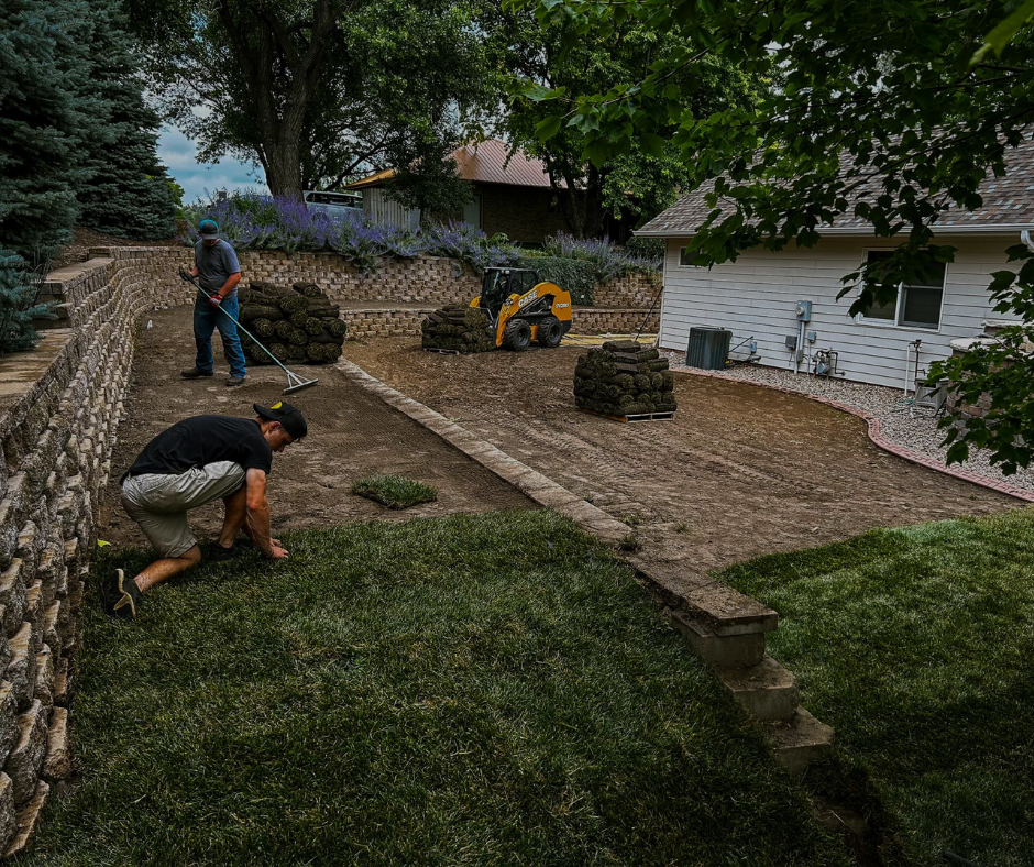 Men laying sod in a backyard with retaining walls, a small skid steer, and a house.