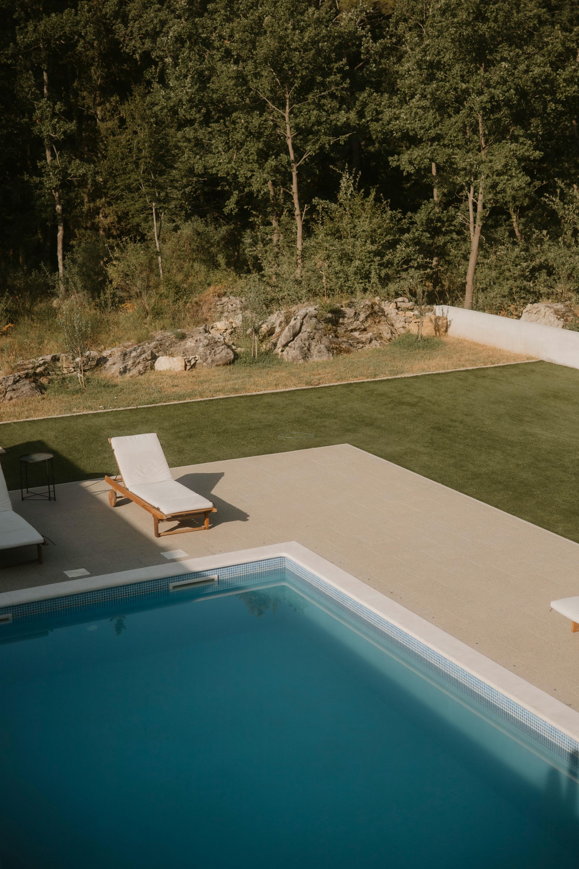 Poolside scene: Blue pool, lounge chairs, green lawn, trees in background.