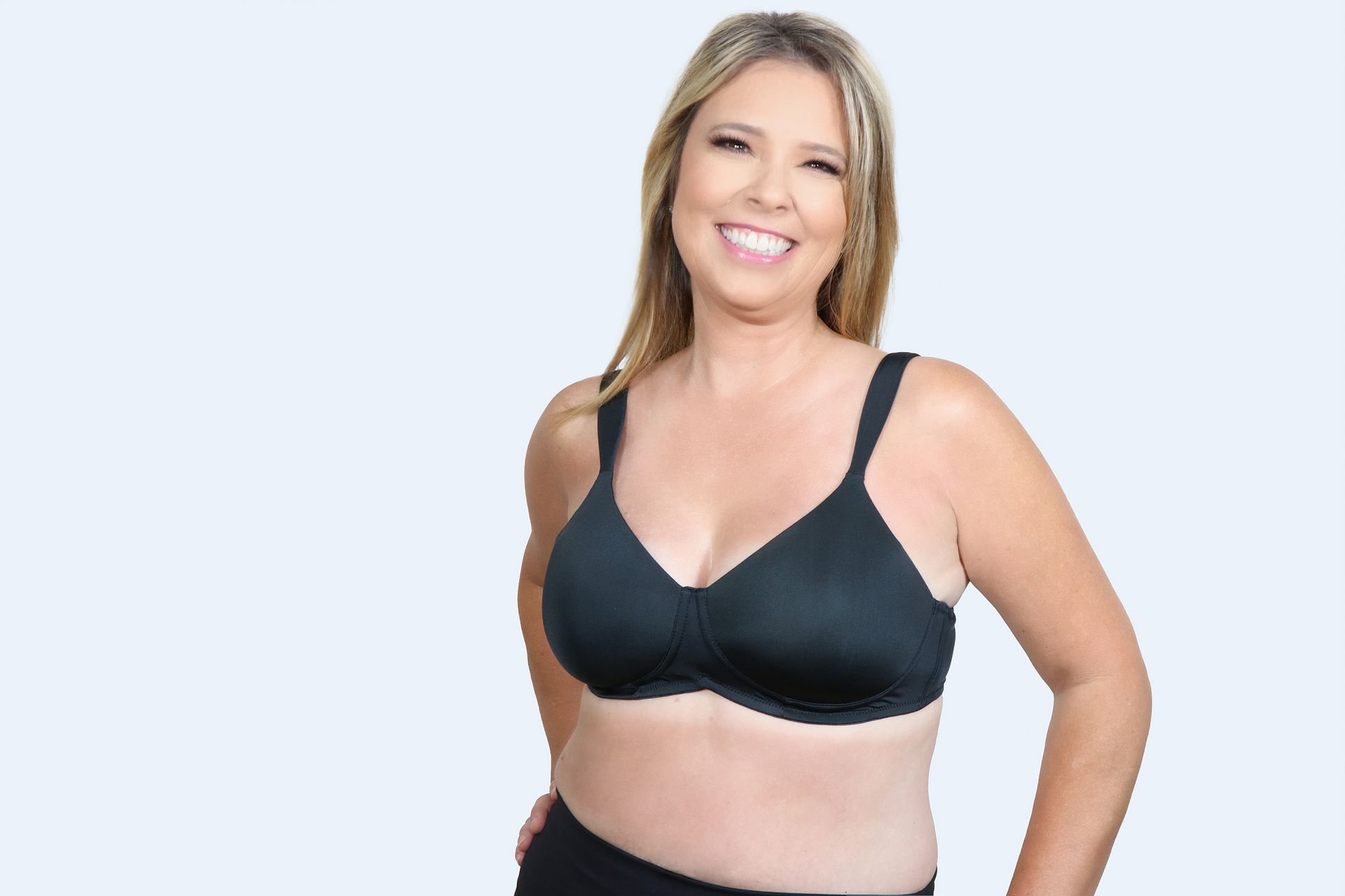 Woman in black bra smiling, standing against a light blue background.
