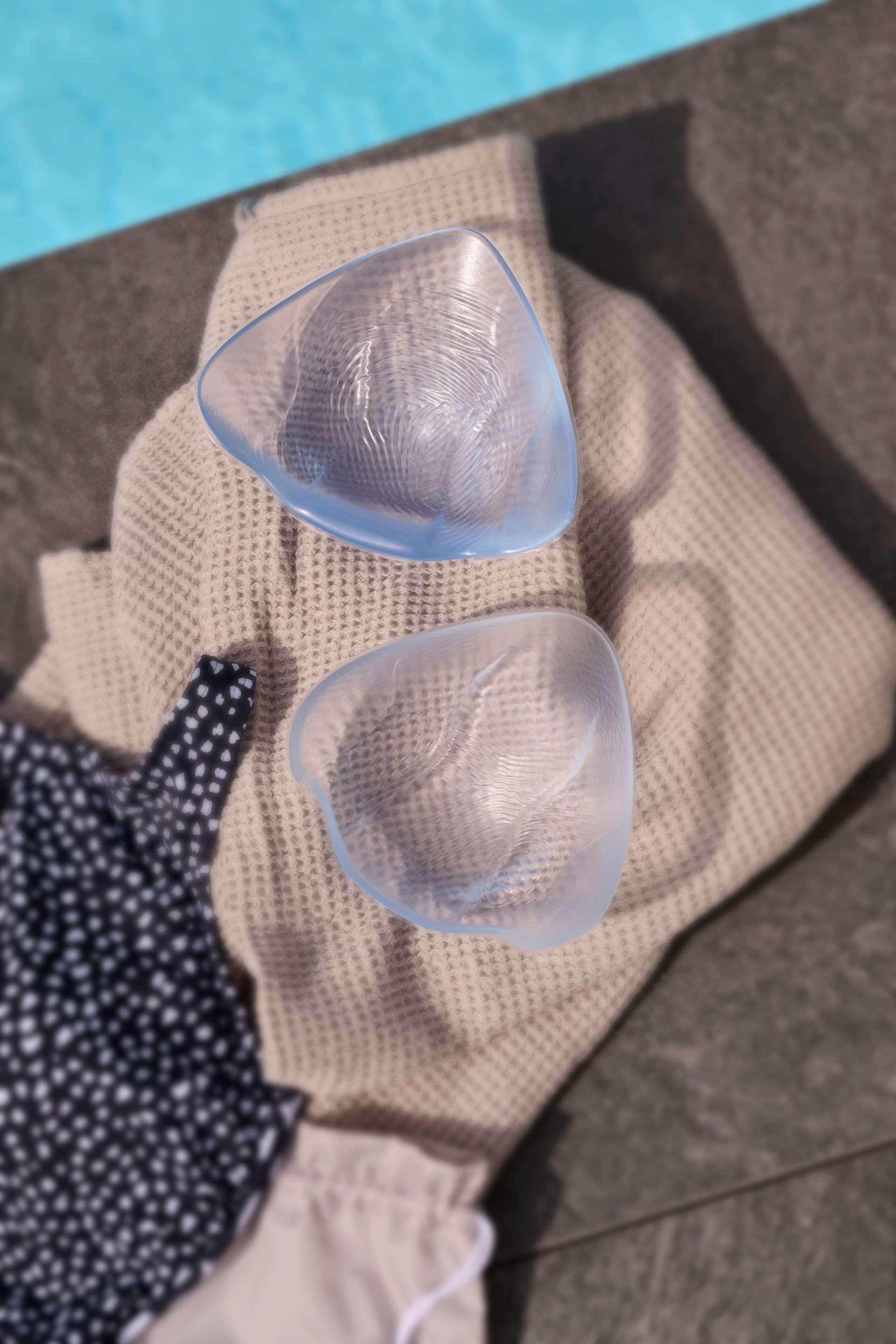 Two clear silicone bra inserts on a beige cloth next to a pool and swimsuit.