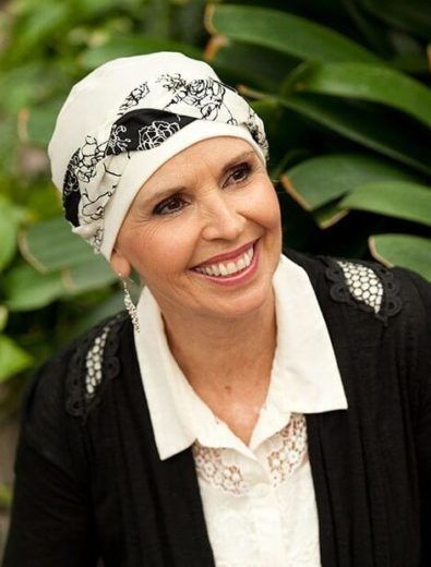 Woman with a patterned headscarf smiling, wearing a black jacket over a white blouse, and dangling earrings.
