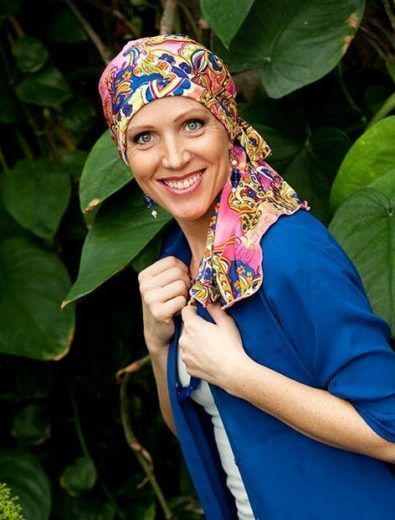 Woman wearing a colorful headscarf smiles, posing in front of lush green foliage, wearing a blue jacket.