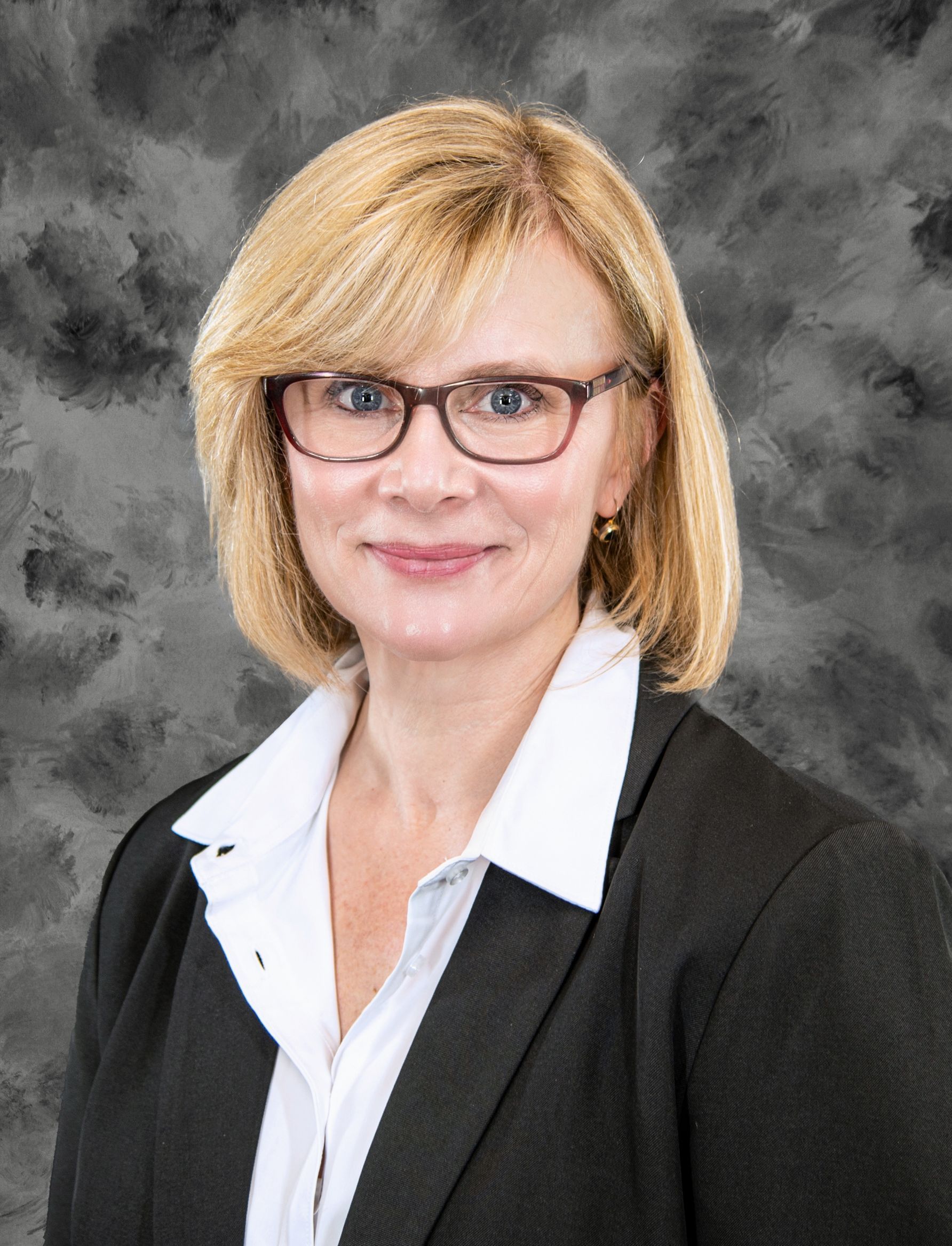 Woman with blonde hair, glasses, and a blazer smiling in front of a gray backdrop.