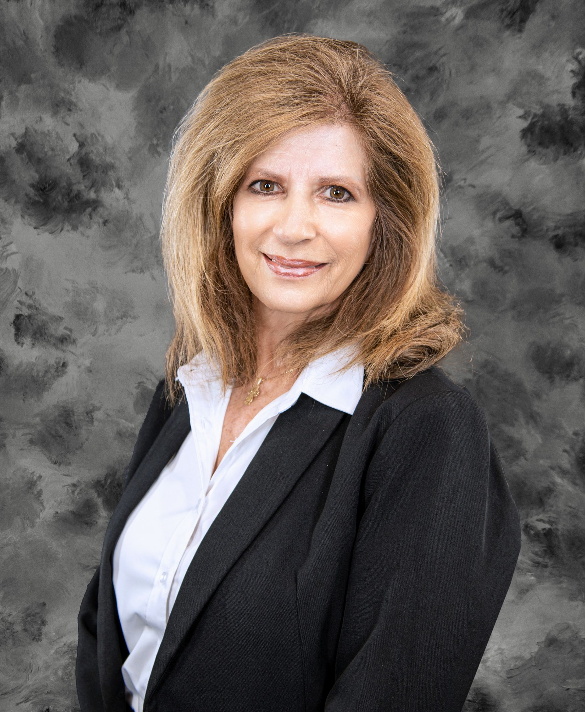 Woman with shoulder-length blonde hair in black blazer and white shirt, smiling. Gray background.