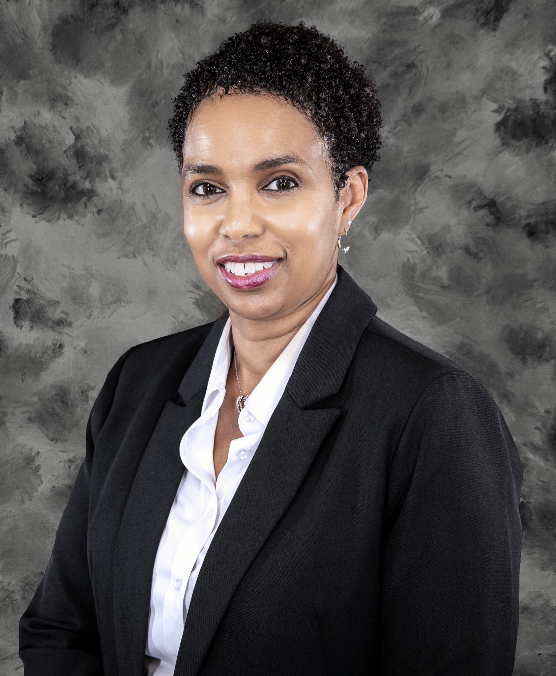 Woman in black blazer and white shirt smiles, against a gray background.