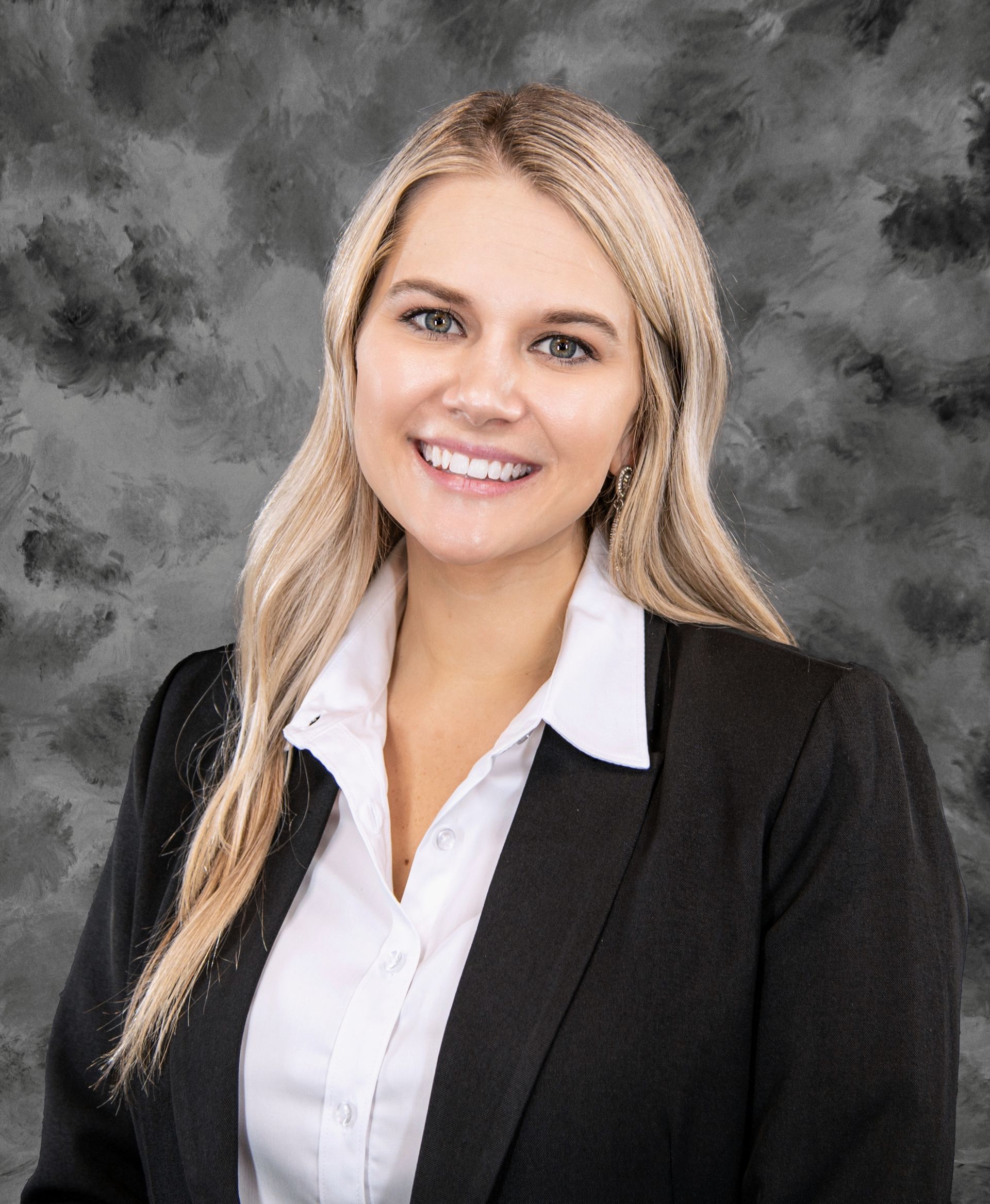 Blonde woman smiles wearing a white shirt and black blazer against a cloudy gray background.