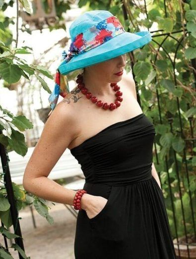 Woman in a black dress and blue hat with a floral scarf, red necklace and bracelet; outdoors.