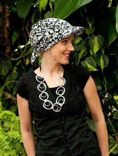 Woman in a patterned hat, black shirt, and silver necklace, smiling in a garden.