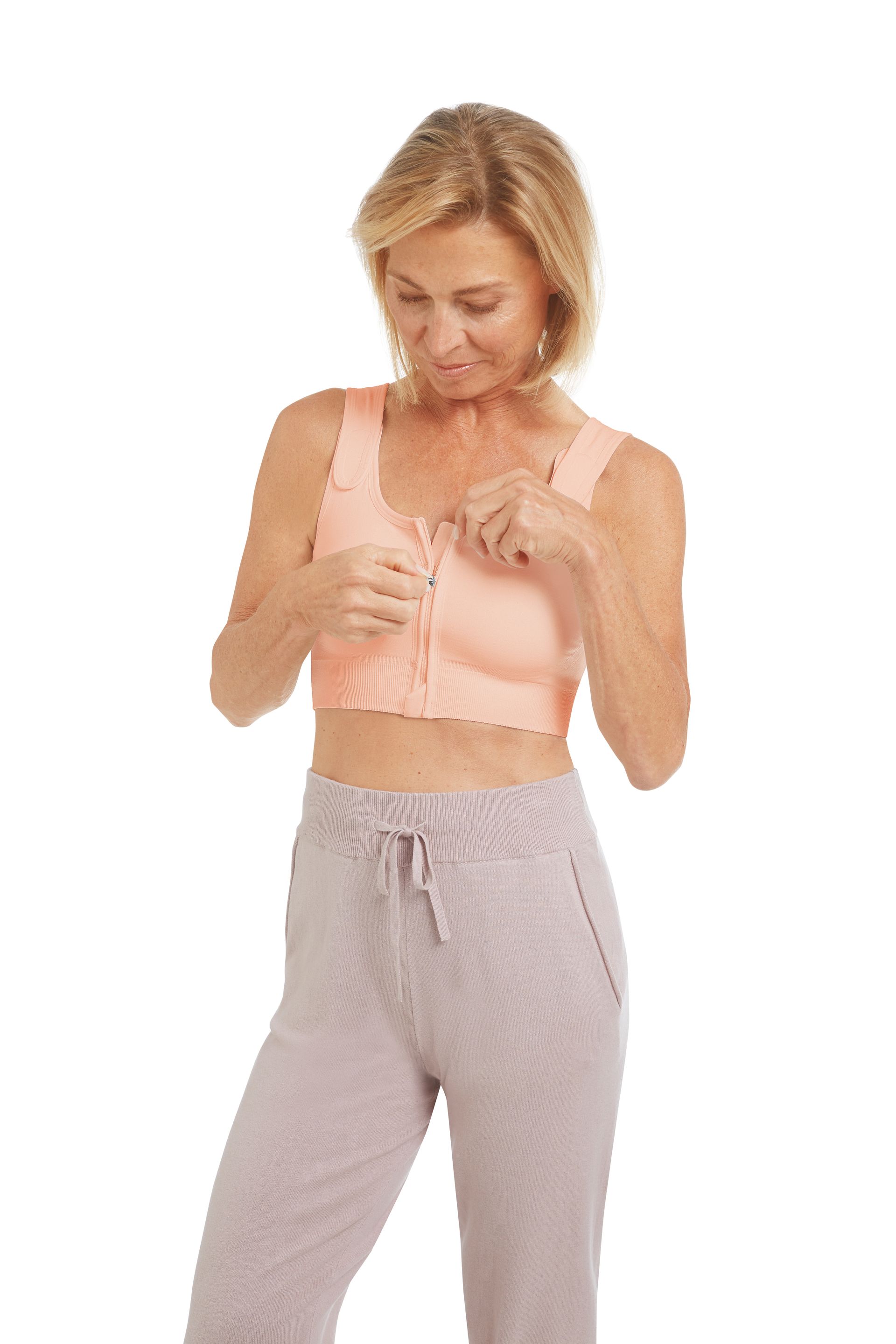 Woman wearing a front-closure peach-colored bra, adjusting the closure. She wears gray sweatpants.