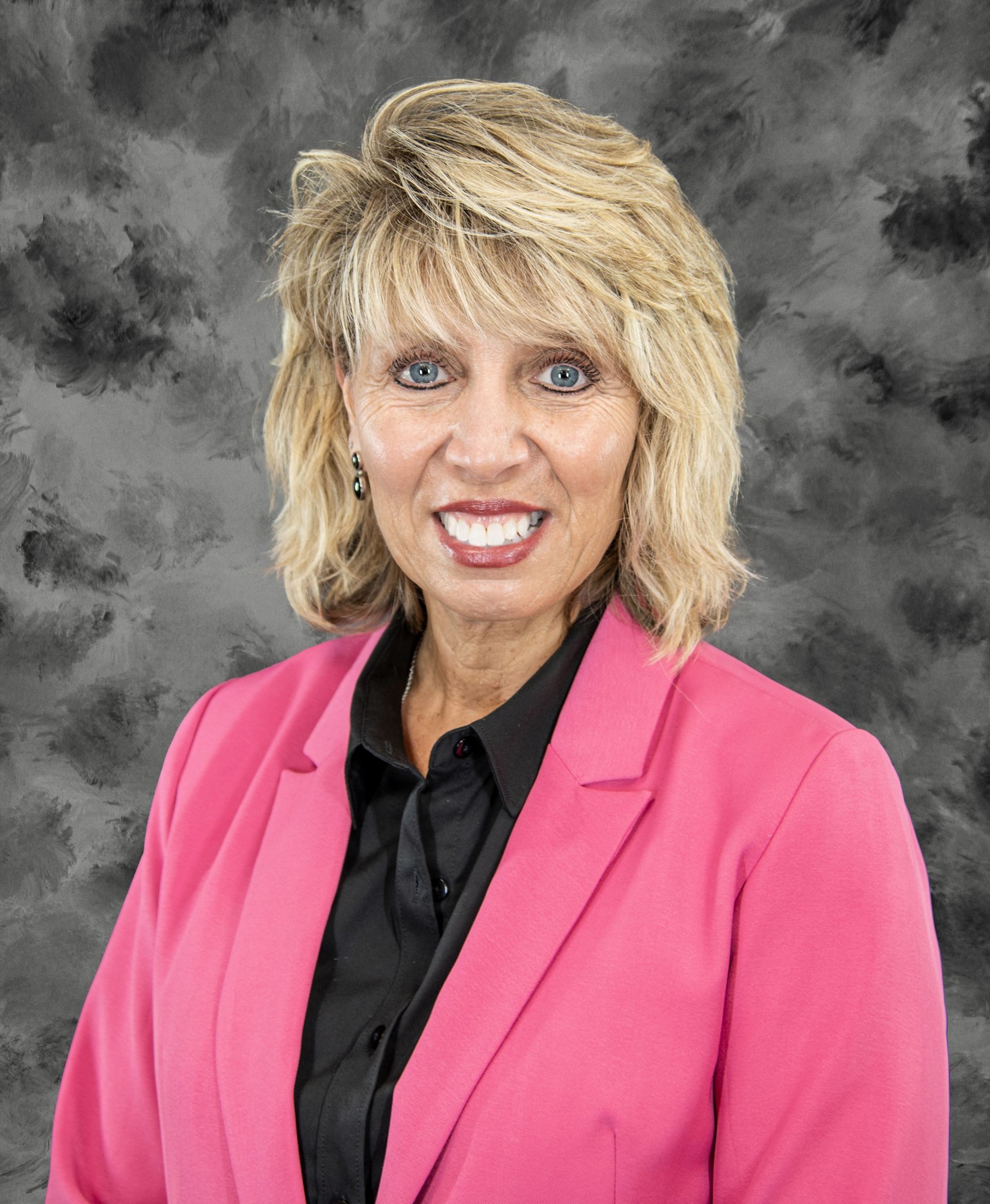 Woman with blonde hair wearing a pink blazer, black shirt, smiling, studio background.