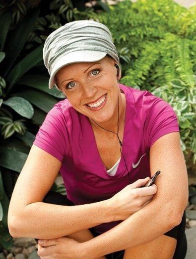 Woman smiling, wearing a cap and pink shirt, holding a device, sitting outdoors with greenery.
