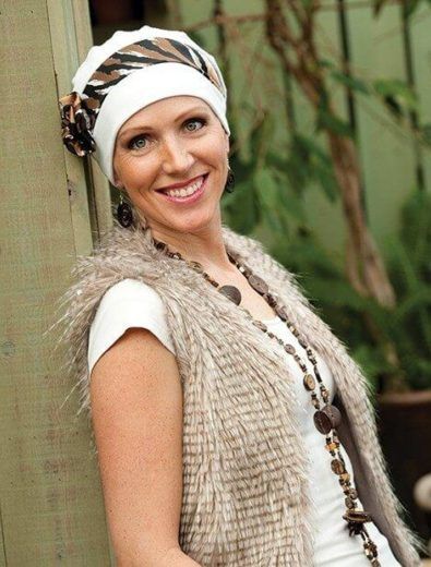 Woman smiling, wearing a white hat with a patterned band, faux fur vest, and wooden beaded necklace.