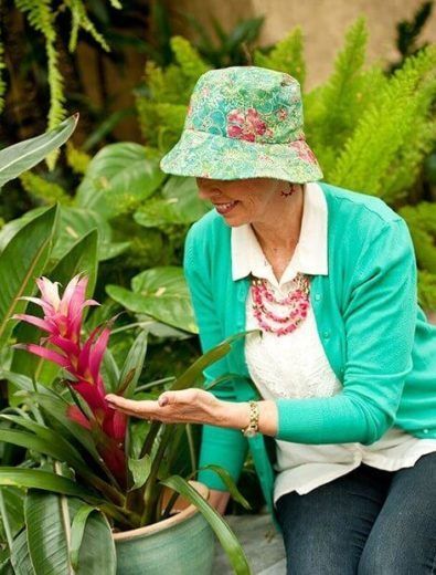 Woman in green hat and cardigan touches a pink flower in a pot, garden setting.